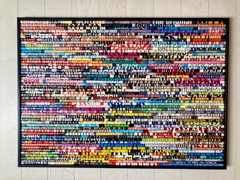 May include: A framed artwork made of colourful magazine strips, each with different text printed on it. The strips are arranged horizontally, creating a layered effect. The frame is black and the background is white.