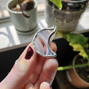 May include: A crescent moon pendant featuring a translucent, light blue-grey banded stone encased in a silver-toned metal frame. The pendant is attached to a delicate silver-toned chain, showcasing a smooth, polished finish on the stone surface.