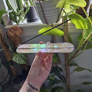 May include: An elongated, rectangular incense holder crafted from iridescent, pearlescent glass with silver-toned metal edges. The holder features a small metal coil at one end designed to securely hold a single, thin black incense stick upright.