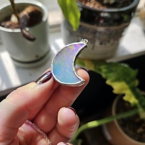 May include: A crescent moon pendant featuring an iridescent, opalescent surface encased in a silver-toned metal frame. The pendant has a smooth, curved shape and a reflective finish that displays shifting colors of blue, pink, and white light.