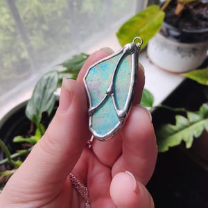 May include: A pendant featuring an iridescent, light blue, semi-translucent piece of glass encased in a silver-toned metal frame with a soldered design. The pendant has a small metal loop at the top for a chain and displays a subtle, shimmering texture.
