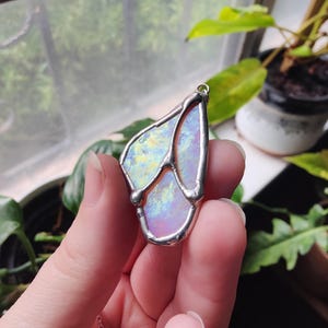 May include: A pendant featuring an iridescent, semi-translucent, light-colored stone encased in a silver-toned metal frame with a soldered design. The piece has a small metal loop at the top for attachment to a thin, delicate silver-toned chain.