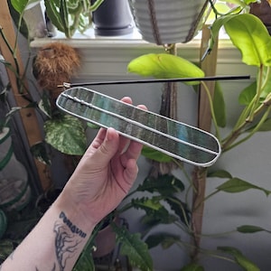 May include: A long, rectangular incense burner made of dark, iridescent stained glass with silver-toned metal solder edges. The burner features a small metal clip at one end, currently holding a single, thin, black incense stick horizontally.