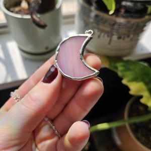 May include: A crescent moon pendant crafted from translucent light pink stained glass with a silver-toned metal solder border. The moon shape features a smooth, curved edge and a small metal loop at the top for attachment to a chain or cord.