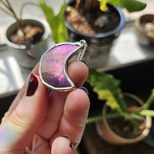 May include: A crescent moon pendant featuring a vibrant purple and pink iridescent surface encased in a silver-toned metal frame. The pendant is attached to a delicate silver-toned chain, showcasing a smooth, reflective finish on the moon shape.