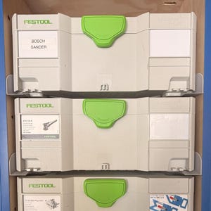 Könnte beinhalten: Drei Festool Werkzeugkisten, die auf einem Regal gestapelt sind. Die Kisten sind hellgrau mit grünen Griffen und weißen Etiketten. Auf der obersten Kiste steht "BOSCH SANDER". Auf der mittleren Kiste steht "ETS 125 R". Auf der unteren Kiste steht "TS 55 REQ-Plus USA".