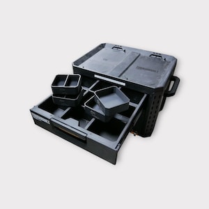 Puede incluir: Caja de almacenamiento de plástico negro con un cajón extraíble y varios compartimentos extraíbles. La caja tiene la palabra "Toughvault" impresa en la parte delantera.