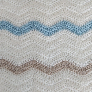 May include: A crocheted blanket featuring a repeating chevron pattern. The design consists of horizontal wavy stripes in alternating colours of off-white, light beige, and soft sky blue, created with a consistent stitch texture throughout the fabric.
