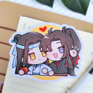 May include: A sticker depicting two cartoon figures with large eyes and smiling faces. One character is in a white and blue outfit, holding a red apple. The other is in a black and red outfit, winking and waving. A red heart is above them.