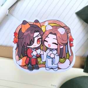 May include: A sticker depicting two anime-style characters. One is in red with fox features, the other in white with bear features. They are holding hands. The background is light brown with red flowers.