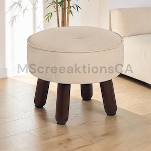 May include: A round, cream-colored upholstered ottoman featuring a central button tuft on the seat and four dark brown cylindrical wooden legs. The text MScreeaktionsCA is overlaid across the center of the ottoman in a light gray font.
