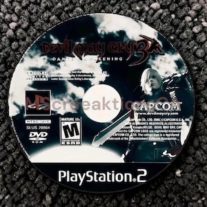Könnte beinhalten: Eine kreisförmige PlayStation 2-Spiel-Disc für Devil May Cry 3: Dante's Awakening. Die Disc zeigt eine schwarz-weiße Grafik der Figur Dante, das Capcom-Logo, eine ESRB Mature 17+-Einstufung und den Produktcode SLUS 20964 auf der Oberfläche.