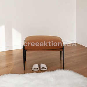 May include: A rectangular ottoman featuring a tan upholstered cushion top supported by a black metal frame with four slender legs. A pair of light-colored slide sandals are positioned directly underneath the center of the ottoman seat.
