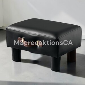 May include: A rectangular black ottoman with a smooth surface and a brown leather-like handle attached to the front side with gold-toned hardware. The piece is supported by four short, cylindrical black legs, and the text MscreeaktionsCA is visible.