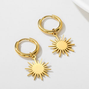 May include: A pair of gold hoop earrings with a dangling sun charm. The sun charm has a starburst design.
