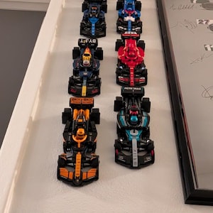 May include: A collection of six miniature Formula 1 race cars arranged in two parallel rows. The models feature various team liveries including orange, dark blue, red, and teal accents, each with detailed cockpits, spoilers, and racing tires.