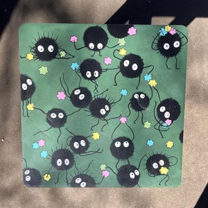 May include: A square, green surface is covered with cartoon soot sprites, each with a fluffy black body, white eyes, and thin black limbs. The sprites are holding colorful star-shaped objects. The background has a textured appearance.