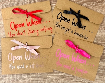 May include: Four brown envelopes with pink and red ribbons tied around them. Each envelope has the text "Open When..." followed by a different phrase: "You don't fancy cooking", "You've got a headache", "You need a hot drink", and "You need a pamper".