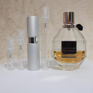 May include: A clear, faceted perfume bottle with a silver cap and a black label that reads "Flowerbomb Viktor & Rolf." Several small, clear spray bottles and a silver atomizer are next to the perfume bottle. The perfume inside the bottle is amber-colored.