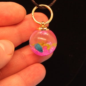 Pode incluir: Um colar de ouro com um pingente de cristal transparente em forma de esfera que contém um peixe azul e glitter rosa. O pingente está suspenso em um cordão preto.