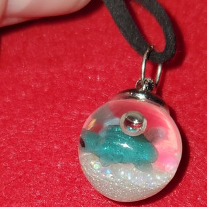 Pode incluir: Um pingente de esfera de vidro transparente com uma figura de tartaruga azul-esverdeada, detalhes prateados e glitter branco. O pingente está preso a um cordão preto.