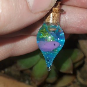 Pode incluir: Um colar com um pingente de garrafa de vidro em forma de lágrima com uma rolha de cortiça. A garrafa está cheia de líquido azul e contém um pequeno objeto rosa e uma folha verde.