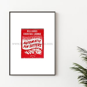 May include: A red matchbook with white text that reads "Billiards Cocktail Lounge Automatic Pin Setters" and a graphic of bowling pins and a bowling ball. The matchbook is framed in a black frame.