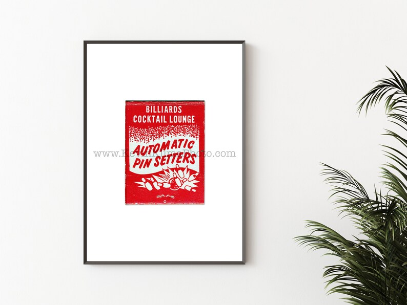 May include: A red matchbook with white text that reads "Billiards Cocktail Lounge Automatic Pin Setters" and a graphic of bowling pins and a bowling ball. The matchbook is framed in a black frame.