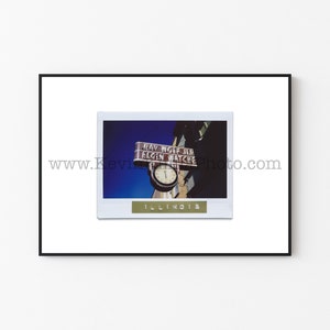 May include: A vintage photograph of a clock with a sign that reads "Ray Wolf Jr. Elgin Watches" in a white frame with the word "Illinois" printed below the image.