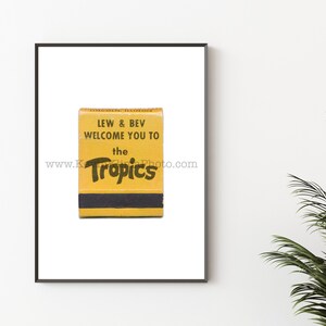 Puede incluir: Una impresión enmarcada con una caja de cerillas vintage. La caja es amarilla y negra, con el texto "LEW & BEV WELCOME YOU TO the Tropics" en una fuente retro. La impresión está sobre un fondo blanco.