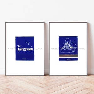 Op de afbeelding: Twee ingelijste prints op een witte muur. De print aan de linkerkant is blauw met witte tekst die "The Rusty Scupper" luidt. De print aan de rechterkant is blauw met een witte omtrek van een schip.