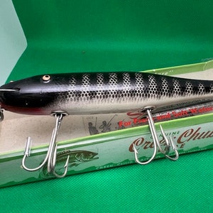 Puede incluir: Un señuelo de pesca negro y blanco con vientre rojo y anzuelos plateados. El señuelo está en su caja original, que es verde y blanca con el texto "Genuine Creek Chub" impreso en ella. La caja también tiene el texto "Para peces de agua dulce y salada".