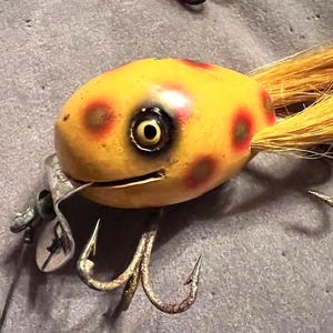 May include: A vintage fishing lure with a yellow body, red spots, and a yellow tail. The lure has a large eye, a metal lip, and two treble hooks. This antique fishing tackle is designed to attract fish.