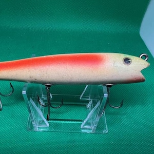 Puede incluir: Un señuelo de pesca de madera con un esquema de pintura rojo y blanco. El señuelo tiene tres anzuelos triples y una boca grande.