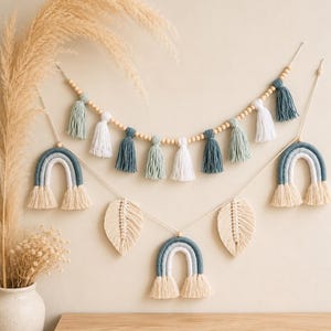 Puede incluir: Un conjunto de decoración de pared con una guirnalda de cuentas de madera y borlas de hilo en azul, verde salvia y blanco. Debajo, dos hojas de macramé y tres arcos de arcoíris en tonos azules y blancos cuelgan de cordones blancos.