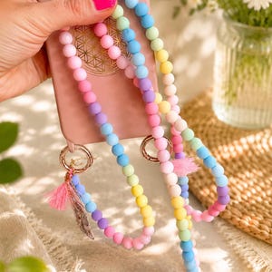 May include: A matte pink smartphone case featuring a gold geometric flower of life emblem on the back. Attached to the case is a long, multi-coloured beaded strap made of round silicone beads in pastel shades of yellow, pink, blue, purple, and green.