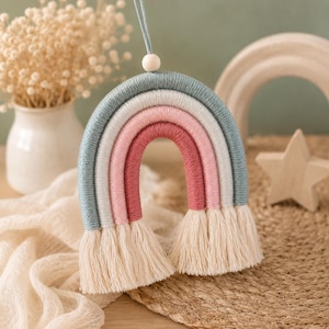 Op de afbeelding: Een decoratieve wandhanger in de vorm van een regenboog, opgebouwd uit vier concentrische bogen omwikkeld met garen in de kleuren stofblauw, wit, lichtroze en donkerroze. De uiteinden hebben crèmekleurige franjes en een houten kraal bovenaan.