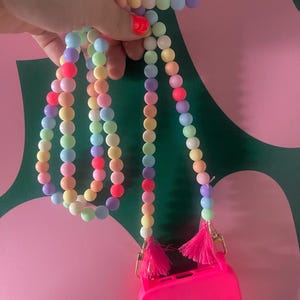 Könnte beinhalten: Eine farbenfrohe, mit Perlen besetzte Handykette in Pastelltönen von Rosa, Gelb, Blau und Grün. Der Riemen ist an einer leuchtend pinkfarbenen Handyhülle mit goldfarbenen Verschlüssen und rosa Quasten befestigt. Die Perlen sind rund und glatt.
