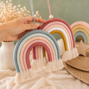 May include: Two hanging rainbow decorations made of wrapped yarn in muted tones of dusty rose, sage green, mustard yellow, and white. Each arch features a fringe of cream-colored cotton cord at the base and a single wooden bead on the hanging string.