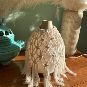 Könnte beinhalten: Ein weißer Keramikdiffusor mit einer cremefarbenen Makramee-Hülle und Fransen. Der Diffusor steht auf einem Holztisch.