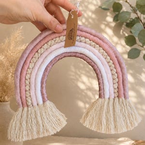 Peut inclure: Une décoration murale arc-en-ciel en macramé composée de cinq couches arquées dans des tons vieux rose, crème et cuivre scintillant. L'objet comporte une étiquette en papier brun avec un logo arc-en-ciel et du texte, finie par des franges crème.