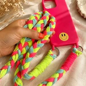 Op de afbeelding: Een felroze telefoonhoesje met een geel smiley-icoon. Het hoesje is bevestigd aan een lange, dikke, gevlochten riem gemaakt van stroken stof in neongeel, felroze en lichtblauw, verbonden aan het hoesje met zilverkleurige metalen ringen.