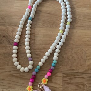 Può includere: Un laccio per telefono a perline bianche con perline colorate e ciondoli a forma di fiore. Il laccio ha una chiusura dorata e un fiocco rosa.