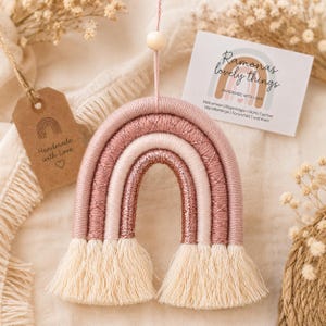 Op de afbeelding: Een hangende regenboogdecoratie gemaakt van gewikkeld garen in tinten oudroze, lichtroze en metallic koper, met een centrale houten kraal en crèmekleurige franjes aan beide uiteinden. Een bruin label leest Handmade with Love en een kaartje Ramonas lovely things.