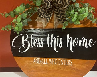May include: A round wooden sign with a black and brown wood grain pattern. The sign has a black painted section with white lettering that reads "Bless this home and all who enters". The sign is decorated with a burlap bow with black and white chevron pattern and two sprigs of green foliage.