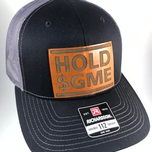 May include: Black trucker hat with a gray mesh back and a brown leather patch. The patch features the words "HOLD $GME" in a bold, embossed font. The hat is a Richardson 112, one size fits all.