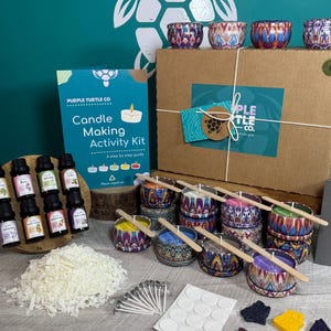 May include: A candle making activity kit by Purple Turtle Co. includes a cardboard box, a step-by-step guide, eight essential oil bottles, a pile of white wax flakes, metal candle tins with patterned designs, wooden wicks, adhesive dots, and coloured wax chunks.