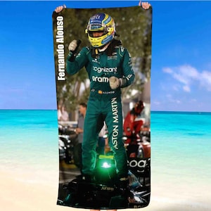 Pode incluir: Uma toalha de praia com um piloto de Fórmula 1 em fato e capacete verdes. A toalha tem o texto "Fernando Alonso" e "Aston Martin" impresso verticalmente. O fundo mostra uma praia com céu azul e oceano.