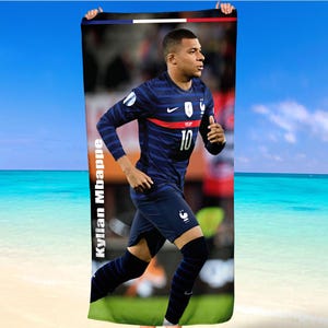 Peut inclure: Une serviette de plage bleue marine avec une image pleine de Kylian Mbappé en uniforme de l'équipe nationale française. La serviette a des détails blancs et rouges, et le nom du joueur est imprimé verticalement en blanc. Le fond montre une plage et un ciel bleu.