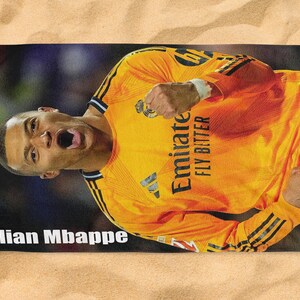 May include: A rectangular beach towel featuring a vibrant image of Kylian Mbapp&eacute; in a yellow soccer jersey. The towel has a red, white, and blue border and the text "Kylian Mbappe". The jersey has the text "Emirate FLY BITTER".